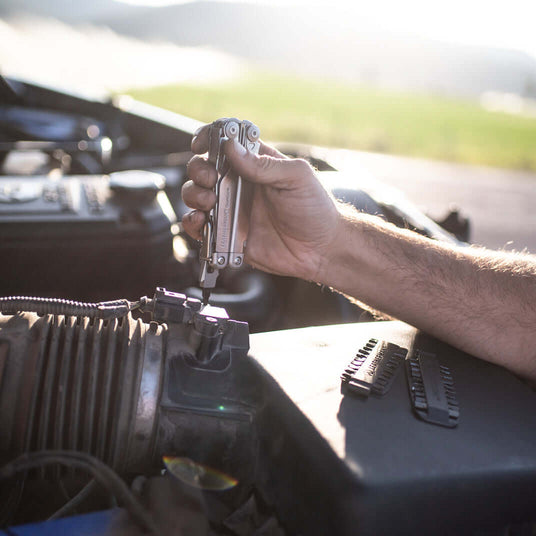 Translation missing: en.Person using a Leatherman Surge® multitool on a car engine in a sunny outdoor setting