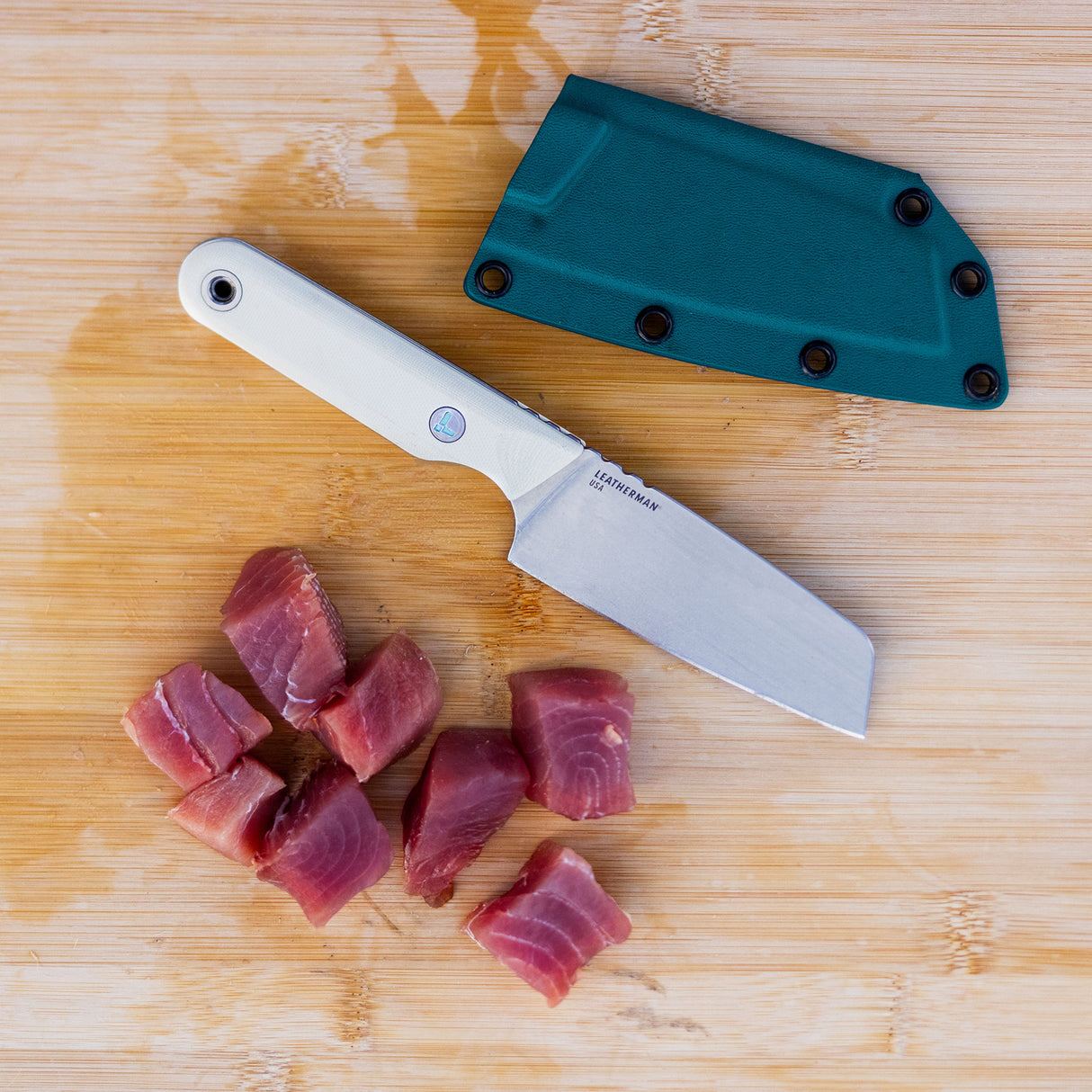 Translation missing: en.A Leatherman Rustle Alpine knife on a wooden cutting board next to a teal sheath and freshly cut chunks of raw fish
