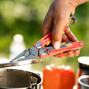 Translation missing: es.Hand using a Guava Leatherman Signal® multitool with pliers to lift a hot metal pot lid while cooking outdoors