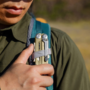Translation missing: es.Person securing a Sandstorm multitool to a backpack shoulder strap with a gray loop outdoors