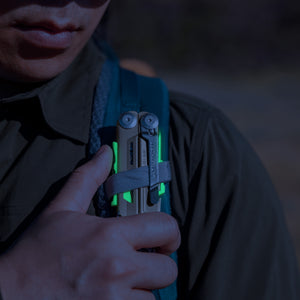 Translation missing: es.Person securing a Sandstorm multitool with neon accents to a backpack strap outdoors