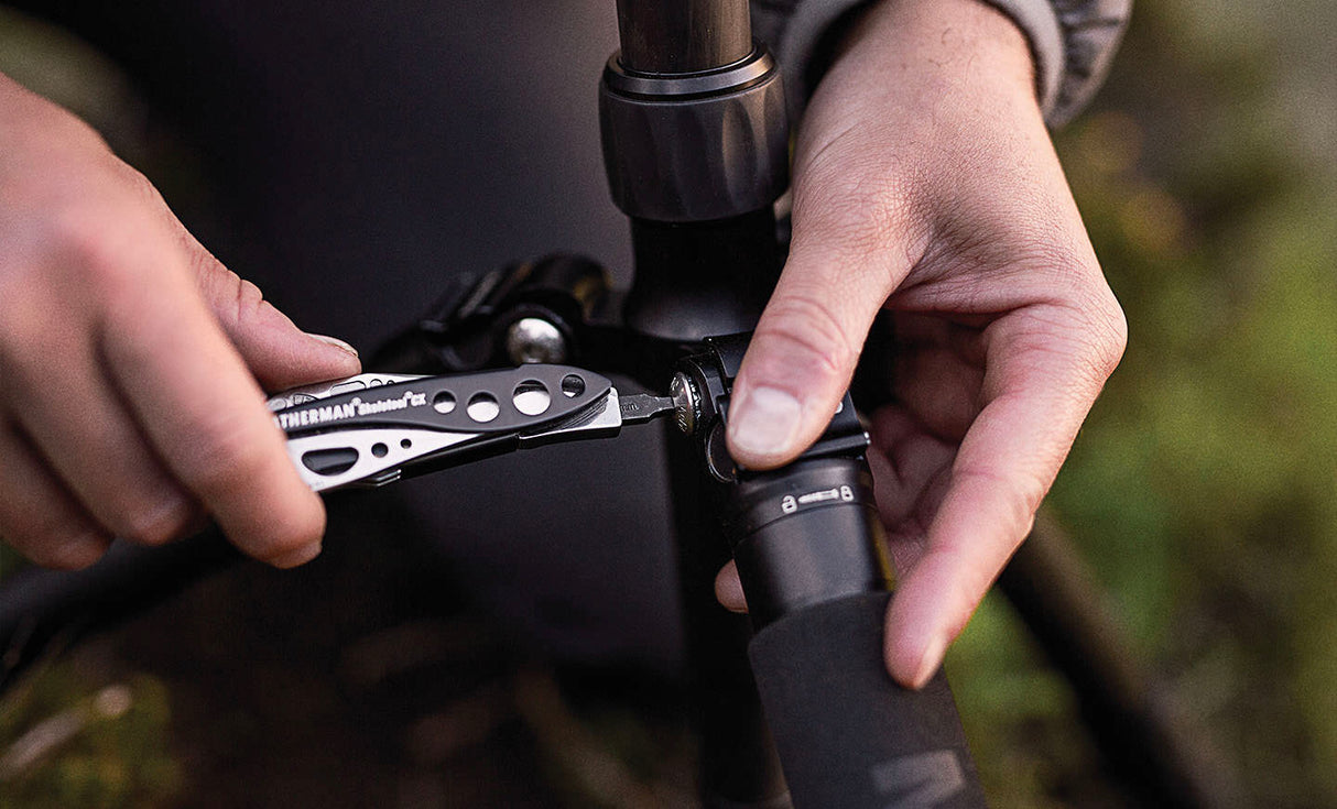 Translation missing: es.Person using a Black &amp; Stainless Leatherman Skeletool® CX multi-tool to adjust a camera tripod in an outdoor setting