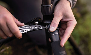 Translation missing: es.Person using a Black &amp; Stainless Leatherman Skeletool® CX multi-tool to adjust a camera tripod in an outdoor setting